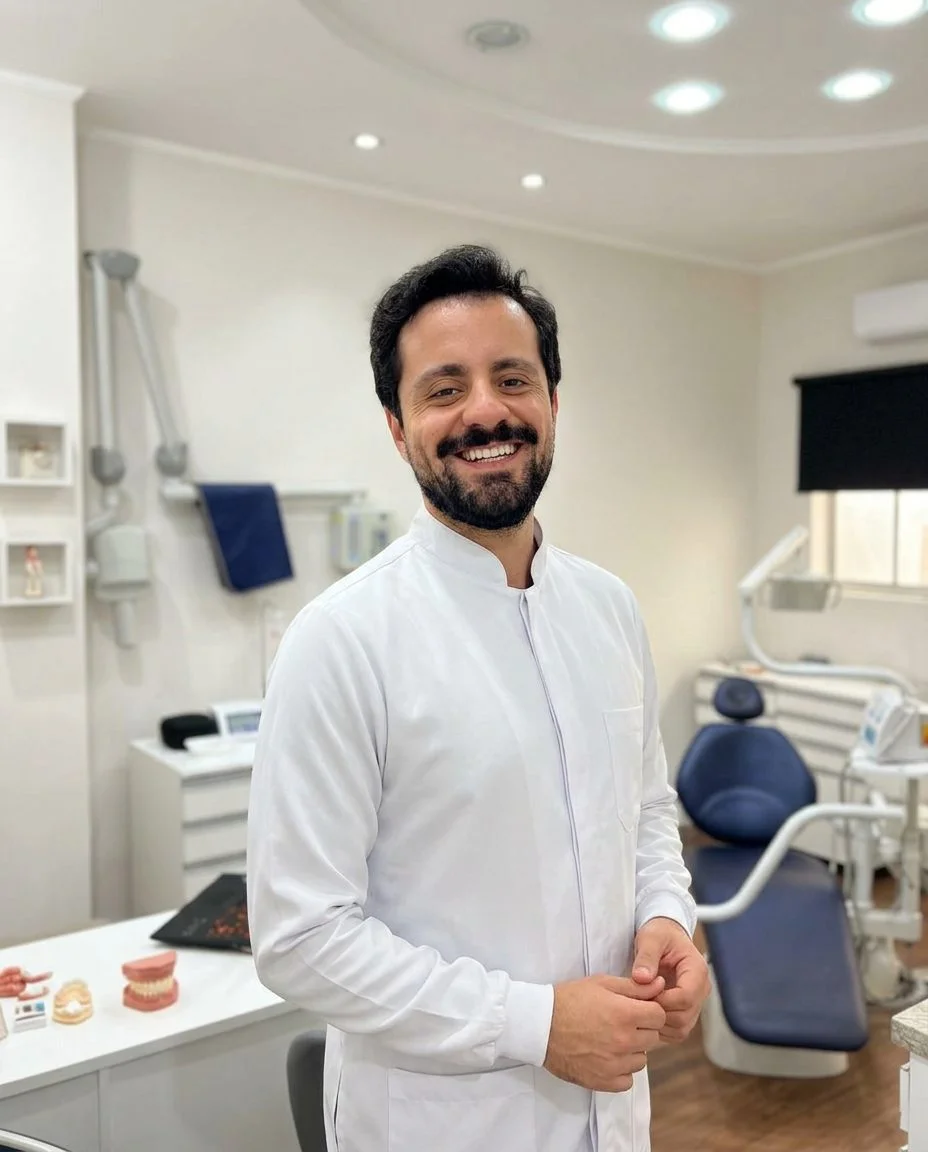 Dr. Alceu Gaiga Junior sorrindo, de jaleco branco, em seu consultório ao lado da cadeira odontológica